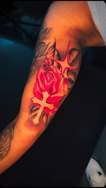 Forearm tattoo of a red rose beside a grey cross